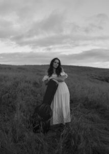 black and white photo of Hayli Carleton leaning against an upright guitar case wearing a white dress in a field of grass.